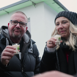 Zwei winterlich angezogene Menschen. Ein Mann und eine Frau, beide mit einem kleinen Glas in der Hand. 