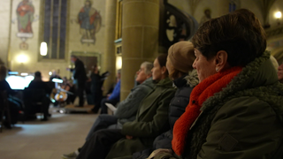 Sonja Faber-Schrecklein sitzt in einer Kirche auf einer Bank. Sie schaut zu einer kleinen Bühne, auf der ein Konzert gegeben wird. 