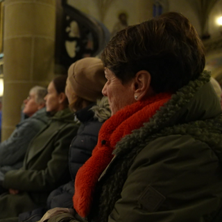 Sonja Faber-Schrecklein sitzt in einer Kirche auf einer Bank. Sie schaut zu einer kleinen Bühne, auf der ein Konzert gegeben wird. 