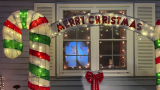 Ein weihnachtlich geschmücktes Fenster. Davor eine große Deko-Zuckerstange und der Schriftzug „Merry Christmas“.