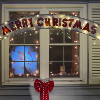 Ein weihnachtlich geschmücktes Fenster. Davor eine große Deko-Zuckerstange und der Schriftzug „Merry Christmas“.