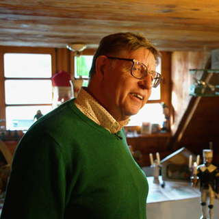 Ein Mann steht in einem Zimmer voller Weihnachtsdekoration. Er trägt einen grünen Pullover und eine Brille. 