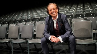 Kabarettist Christoph Sonntag sitzt in einem Raum voller Stühle in der ersten Reihe.
