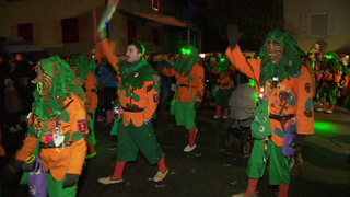 Verkleidete Menschen ziehen in einem bunten Fasnachtsumzug durch das Dorf, umringt von jubelnden Zuschauern.
