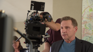 Richy Müller sitzt am Filmset, hinter ihm ist eine professionelle Kamera für die Dreharbeiten zu sehen.