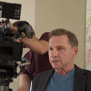 Richy Müller sitzt am Filmset, hinter ihm ist eine professionelle Kamera für die Dreharbeiten zu sehen.