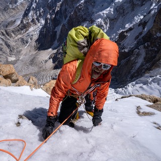 Extrembergsteiger Thomas Huber klettert im Hochgebirge. Er ist mit einem Seil gesichert, trägt eine orangene Jacke, Sonnenbrille und einen gelbgrünen Rocksack.