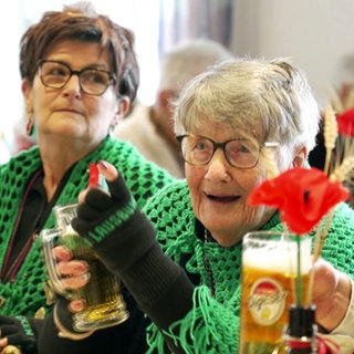 Mit fast 91 Jahren ist Thea Breuling als Lausbühlhexe bei der Fasnet in Ergenzingen aktiv. Sie sitzt mit anderen Narren in einem Saal, trägt einen grünen Umhang und trinkt ein Bier.