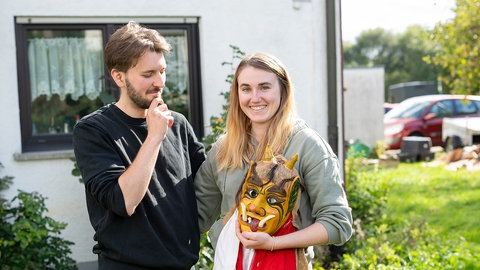Alina Freisler und Pascal Schwarz stehen vor einem Haus. Die beiden machen bei der ARD Serie 'Raus aufs Land' mit. Sie hält eine Maske in der Hand, er schaut fragend darauf.
