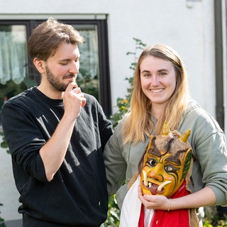 Alina Freisler und Pascal Schwarz stehen vor einem Haus. Die beiden machen bei der ARD Serie 'Raus aufs Land' mit. Sie hält eine Maske in der Hand, er schaut fragend darauf.