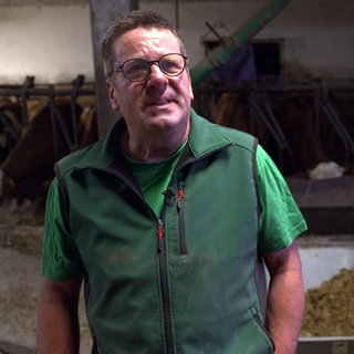 Betriebshelfer Herbert Küpper steht in einem Kuhstall. Hinter ihm sind Kühe und eine Schubkarre mit Mist zu sehen. Er trägt ein grünes T-Shirt und eine grüße Weste.