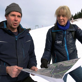 Skiliftbetreiber Matthias Schneider und Andrea Brender stehen im Schnee auf der Skipiste. Sie halten einen Landschaftsplan in der Hand.
