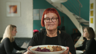 Eine Frau mit roten Haaren und Brille hält einen Teller in der Hand. Im Hintergrund sitzen zwei Frauen an einem Tisch. 