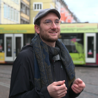 Ein junger Mann trägt eine Schiebermütze und lächelt. Er steht auf einer Straße. Sein name ist Terence Hill.