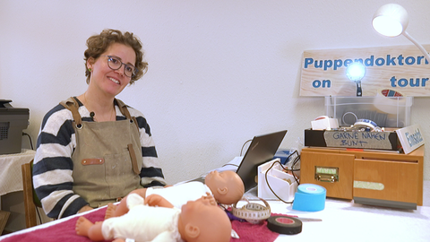 Puppenärztin Anja Müller sitzt an einem Tisch. Vor ihr liegen zwei Puppen, ein Maßband und Nähgarn. 