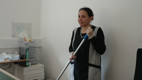 Reinigungskraft Ayse steht in einem Büro. Sie trägt einen Putzkittel und hat Handschuhe an. In den Händen hält sie einen Wischmopp.