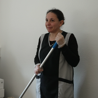 Reinigungskraft Ayse steht in einem Büro. Sie trägt einen Putzkittel und hat Handschuhe an. In den Händen hält sie einen Wischmopp.