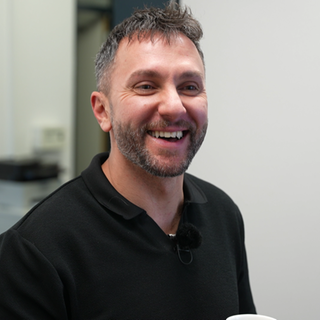 Ein Mann, Hora Soruika, lacht in die Kamera. Er trägt ein schwarzes Shirt.