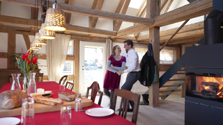 Ein Paar tanzt in einer sanierten Scheune. Im Vordergrund ist ein gedeckter Tisch. Ein Feuer brennt in einem Kamin.