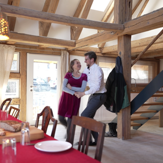 Ein Paar tanzt in einer sanierten Scheune. Im Vordergrund ist ein gedeckter Tisch. Ein Feuer brennt in einem Kamin.