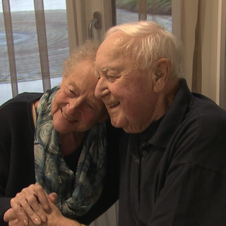 Eine Frau und ein Mann sitzen nebeneinander. Sie sind beide Senioren. Die Frau legt ihren Kopf auf die Schulter des Mannes. Beide lächeln. 