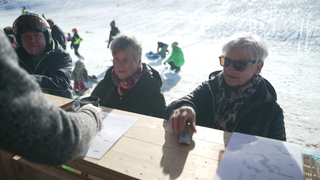 Zwei Frauen stehen an einer Après Ski-Hütte. Im Hintergrund Kinder die rodeln. 