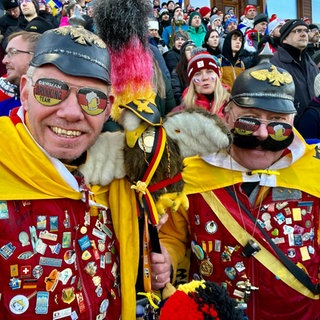 Jörg Himmler und Lars Krause unterstützen die deutschen Teams bei der Winterolympiade. Sie tragen Pickelhauben, Brillen und eine Fahnen mit Deutschland-Farben und eine Jacke mit vielen Broschen.