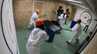 4 Frauen halten Baseballschläger in der Hand und schlagen auf einen Fernseher ein. Sie tragen weiße Overalls und einen orangenen Schutzhelm.