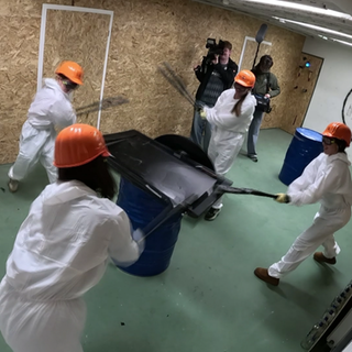 4 Frauen halten Baseballschläger in der Hand und schlagen auf einen Fernseher ein. Sie tragen weiße Overalls und einen orangenen Schutzhelm.