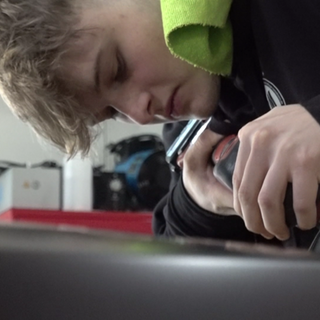 Niklas poliert den Spoiler eines schwarzen Autos. Über seiner Schulter hängt ein grünes Handtuch.