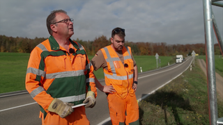 Zwei Männer von der Straßenmeisterei stehen vor einem Straßenschild. Sie tragen orange Arbeitskleidung. 