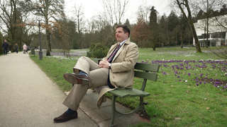 Timo Wolfinger sitzt auf einer Parkbank. Er trägt einen Trenchcoat. 