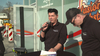 Panajotis Neuert steht an einem Stehtisch und hält ein Mikrofon in seiner Hand. 