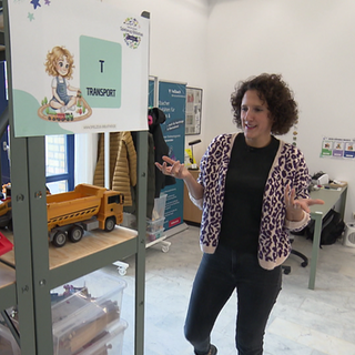 Daniela Bundschuh, eine Frau mit lockigen Haaren, steht in ihrer Spielzeug-Bibliothek. Neben ihr ist ein Regal mit Spielzeug mit der Beschriftung: Transport.