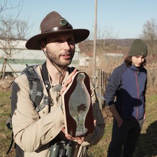 Ein Ranger, steht in einer Gruppe von Menschen. Er hält eine Holztafel hoch, auf der ein Rebhuhn abgebildet ist. 