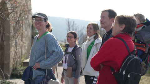 Eine Gruppe von Leuten steht im Sonnenschein draußen. Auf der linken Seite ist verschwommen eine Burgmauer zu sehen. Rechts daneben steht eine Frau der Gruppe, die Richtung Kamera lächelt.