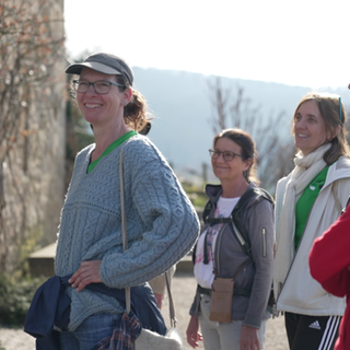 Eine Gruppe von Leuten steht im Sonnenschein draußen. Auf der linken Seite ist verschwommen eine Burgmauer zu sehen. Rechts daneben steht eine Frau der Gruppe, die Richtung Kamera lächelt.