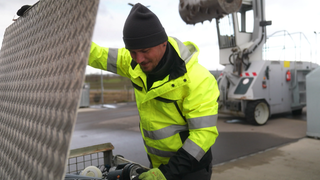 Arno Heinzmann steht in einer neongelben Weste neben einer Kiste voller Elektroschrott.