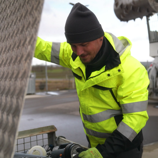 Arno Heinzmann steht in einer neongelben Weste neben einer Kiste voller Elektroschrott.