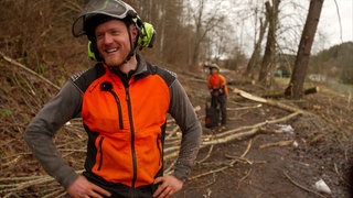 Philipp Klauser stet im Vordergrund und lacht, während er eine orangene Warnweste im Wald trägt.