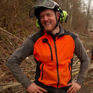 Philipp Klauser stet im Vordergrund und lacht, während er eine orangene Warnweste im Wald trägt.
