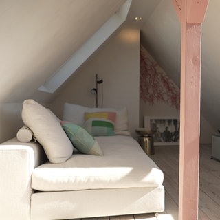 In einem Dachraum steht auf der linken Seite ein weißes Sofa, auf der rechten ein Fernseher. In der Mitte steht ein heller Holzbalken.