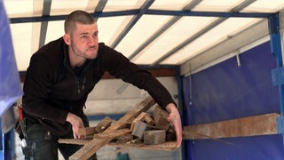 Simon steht in einem Anhänger und trägt einen Haufen Bauschutt in beiden Händen.