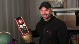 Nico lächelt in die Kamera und hat eine Kuhglocke mit der Schweizer Flagge in der Hand.
