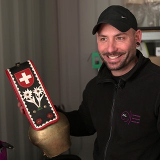Nico lächelt in die Kamera und hat eine Kuhglocke mit der Schweizer Flagge in der Hand.