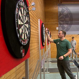 In einer Sporthalle hängen mehrere Dartscheiben, an denen drei Männer spielen. Im Hintergrund steht eine riesige Dartscheibe.