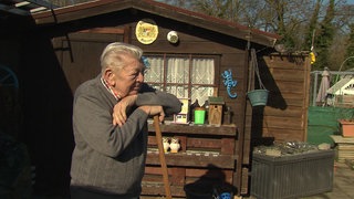 Eckhard Jahrsetz stützt sich auf einer Harke ab und guckt zum rechten Bildrand. Im Hintergrund steht eine braune Gartenhütte.