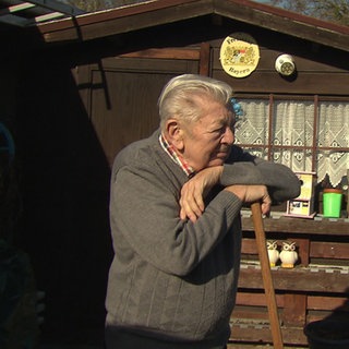 Eckhard Jahrsetz stützt sich auf einer Harke ab und guckt zum rechten Bildrand. Im Hintergrund steht eine braune Gartenhütte.