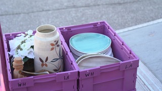 In zwei lila Kisten liegen alte Teller und eine Vase mit Blumenverzierungen.