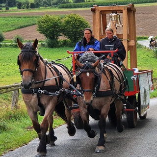 Helga Teichmann und Hubert Wolf fahren mit einer Pferdekutsche auf einer Straße. Auf der Kutsche transportieren sie eine Glocke.